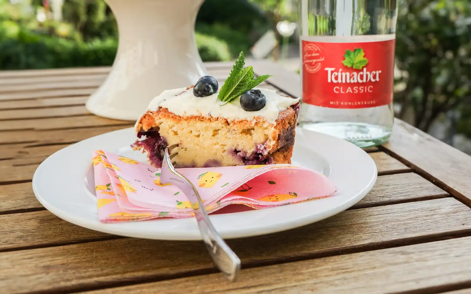 Vorderansicht einer Teinacher 0,75 Liter Mineralwasser Classic Glasflasche und einen Blaubeerkuchen auf einem Teller im Vordergrund