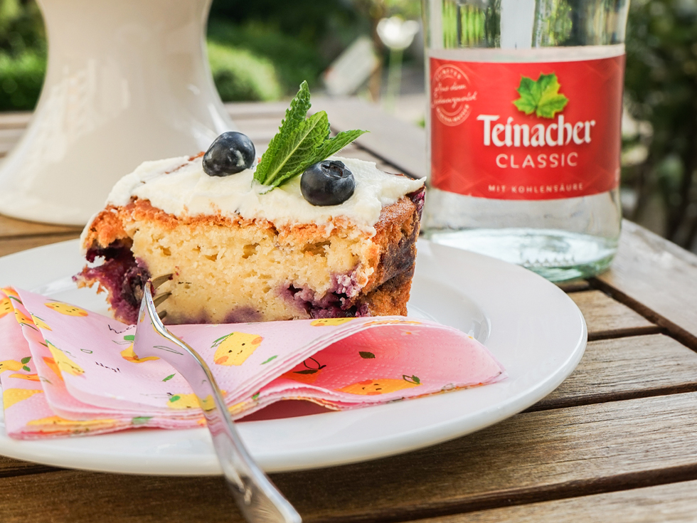 Vorderansicht einer Teinacher 0,75 Liter Mineralwasser Classic Genuss-Flasche mit einem Blaubeerkuchen und Blaubeeren auf einem Teller